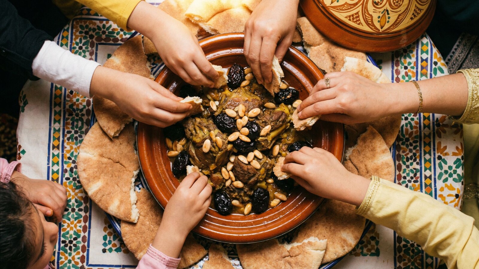 Moroccan communal dining scene showing hands reaching toward a shared tagine with bread around the edges