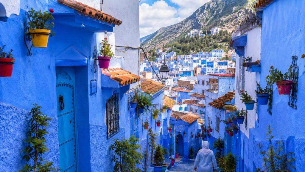 Blue-painted alley in Chefchaouen, Morocco, lined with colorful flowerpots, traditional doors, and views of the Rif Mountains.