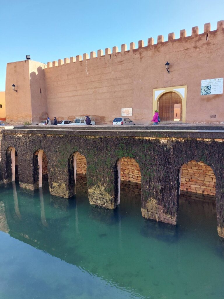 Clay ramparts of Tiznit city with crenellated walls and arched green water moat bridge, a few cars and people visible under a clear blue sky