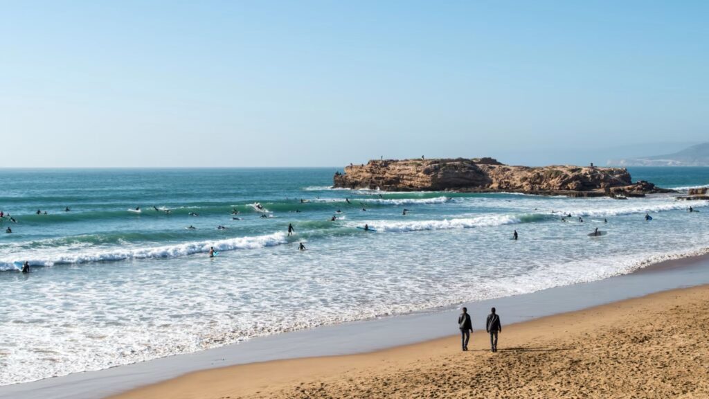 Surfers riding small waves near a rocky outcrop while two people walk along the sandy beach in Tamraght under a clear blue sky.