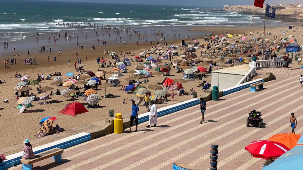 Busy summer day on Aglou Beach with crowds of people relaxing under colorful umbrellas, families playing in the sand, and swimmers enjoying the waves next to a wide seaside promenade