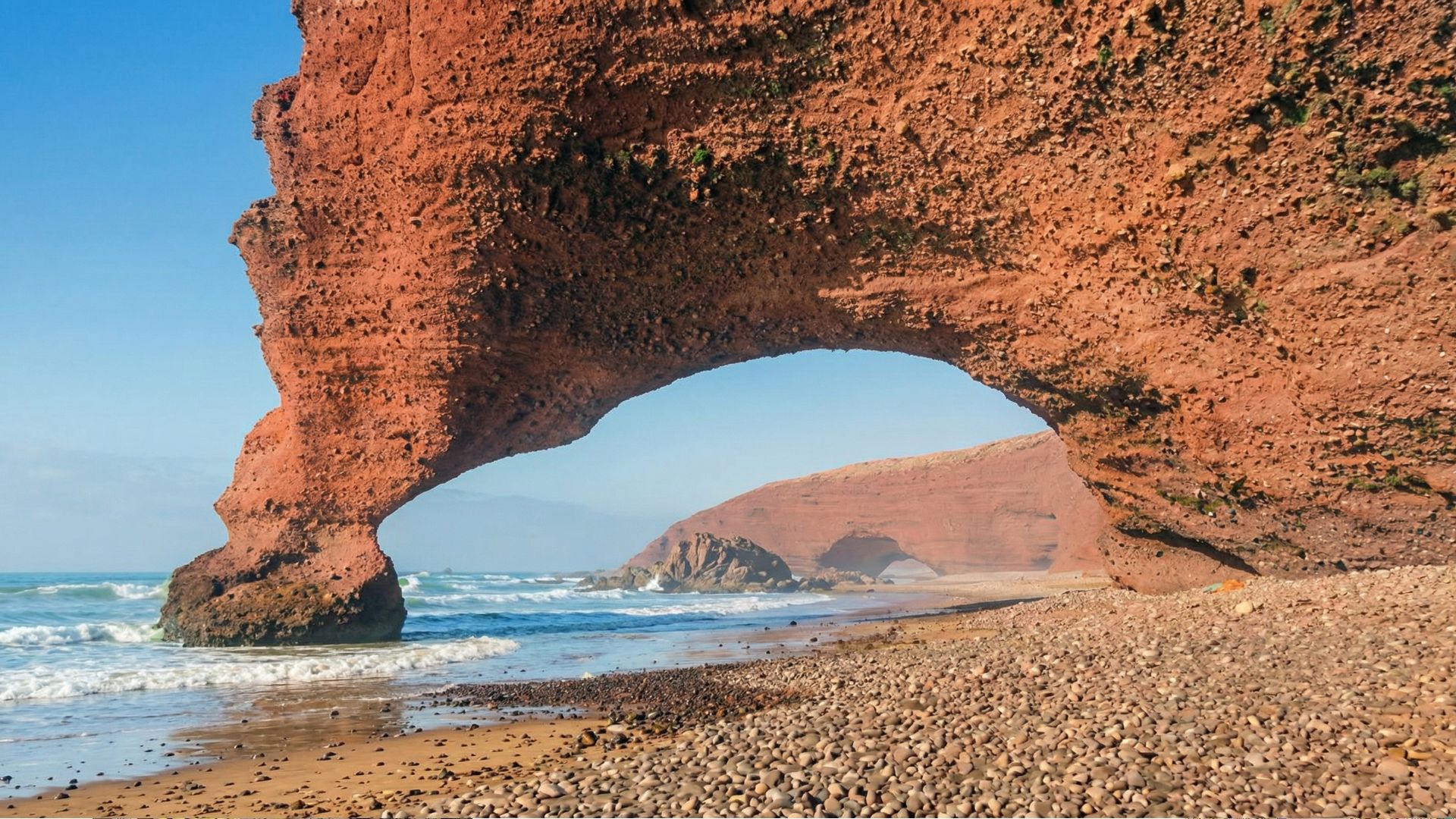 Natural red sandstone arch at Legzira Beach curving over a pebbled shoreline with gentle Atlantic waves and clear blue sky in the background