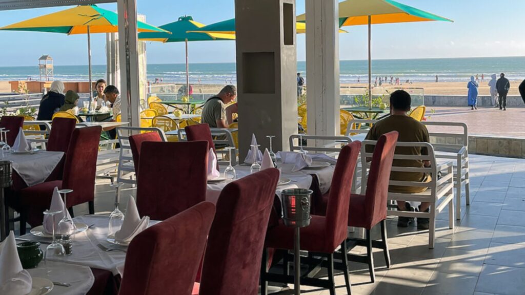Guests dining at an Agadir beachfront restaurant with neatly set tables, red chairs, and yellow-green umbrellas on the terrace overlooking the promenade and Atlantic Ocean