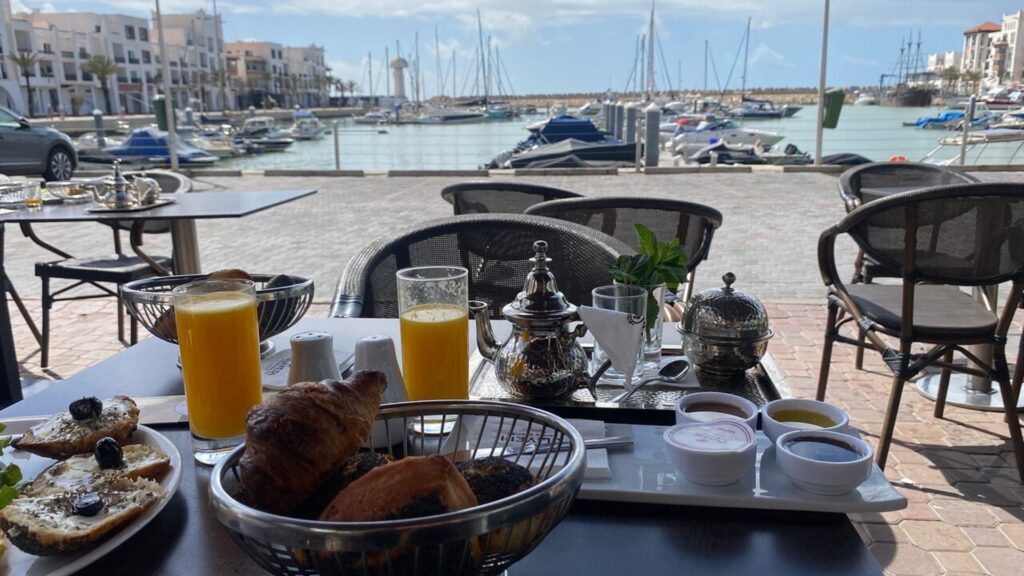 Alt text: Breakfast table at a Marina Agadir restaurant with croissants, Moroccan mint tea, orange juice, and jams, overlooking yachts and white waterfront apartments in the harbor
