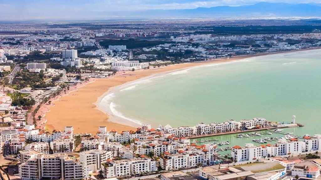 Aerial view of Agadir’s crescent-shaped sandy beach, turquoise Atlantic waters, and white resort buildings lining the marina and coastline in southern Morocco