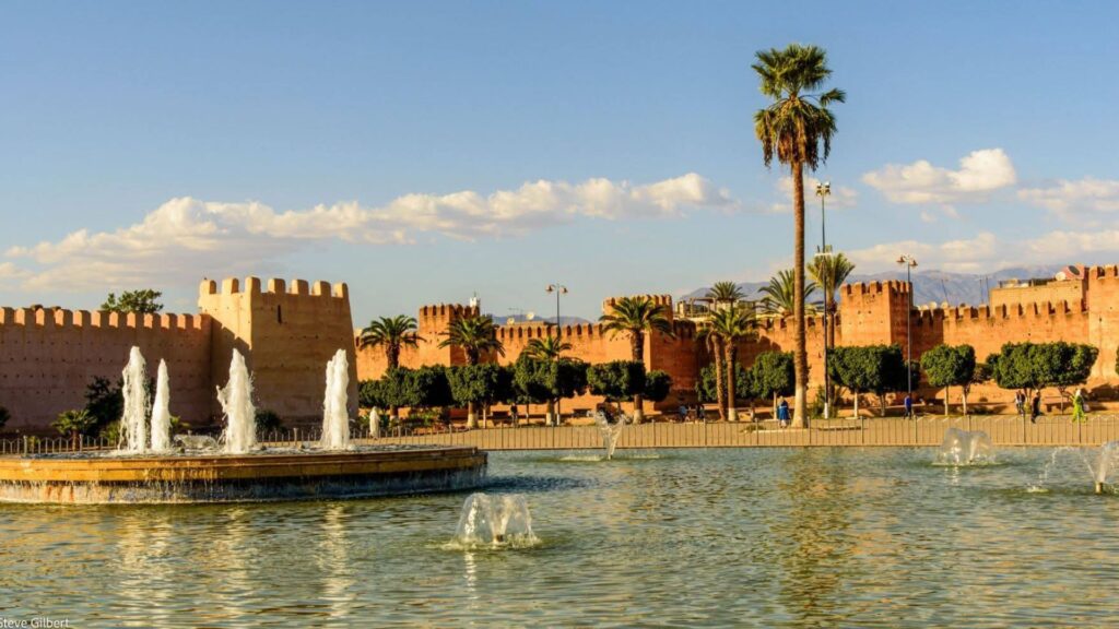 Sunlit ramparts of Taroudant city reflected in a large fountain pool, with palm trees, greenery, and the distant Atlas Mountains under a blue sky with scattered clouds