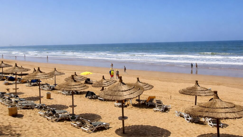 Scenic view of Agadir Beach in Morocco with golden sand, Atlantic waves, straw umbrellas, sun loungers, and people walking along the shoreline under a clear blue sky.