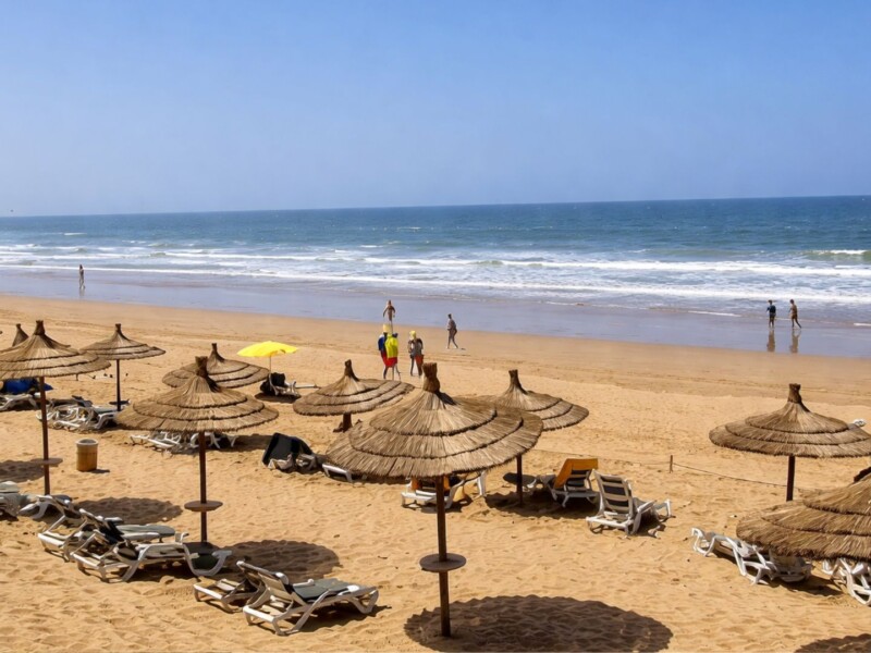 Scenic view of Agadir Beach in Morocco with golden sand, Atlantic waves, straw umbrellas, sun loungers, and people walking along the shoreline under a clear blue sky.