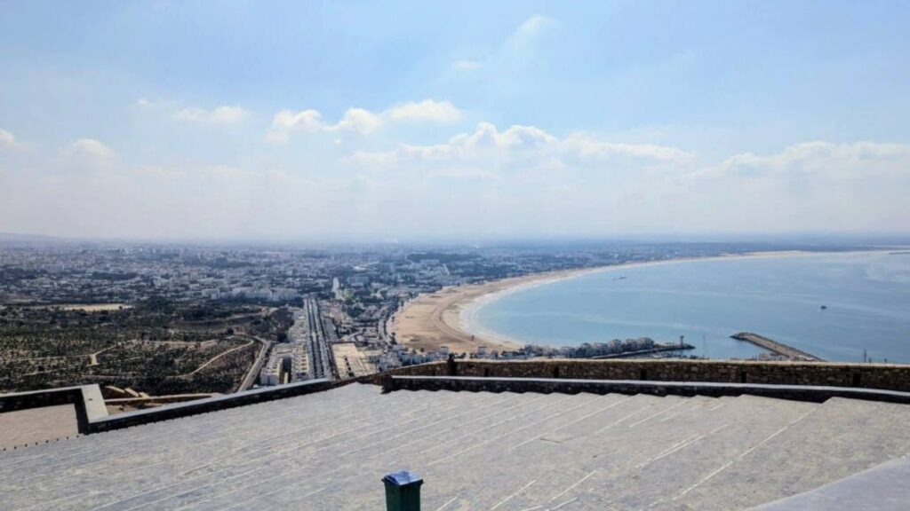 Panoramic view from the hilltop above Agadir, overlooking the long sandy beach, calm Atlantic bay, and sprawling cityscape under a hazy blue sky