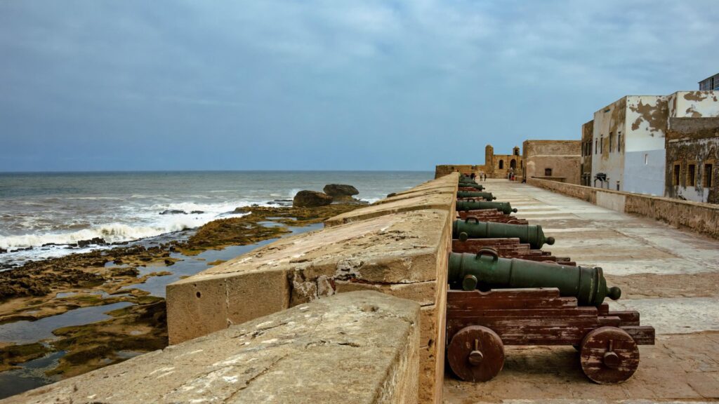 Historic stone ramparts and line of old cannons overlooking the rocky Atlantic coastline in Essaouira, Morocco, under a cloudy blue sky