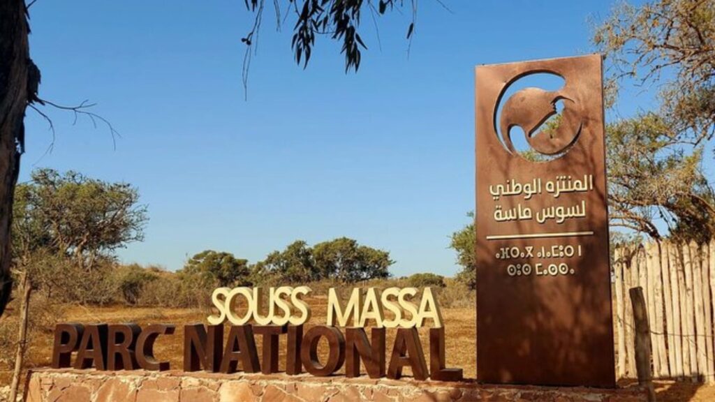 Entrance sign of Souss Massa National Park in southern Morocco, with modern brown lettering in French and Arabic set against a dry savanna landscape and clear blue sky