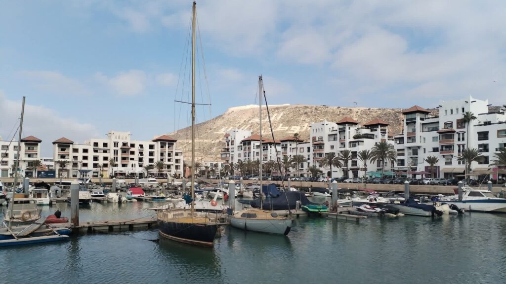 Boats moored in Agadir Marina with white Mediterranean-style apartments, palm trees, and the hillside “Allah, Al Watan, Al Malik” inscription in the background under a partly cloudy sky