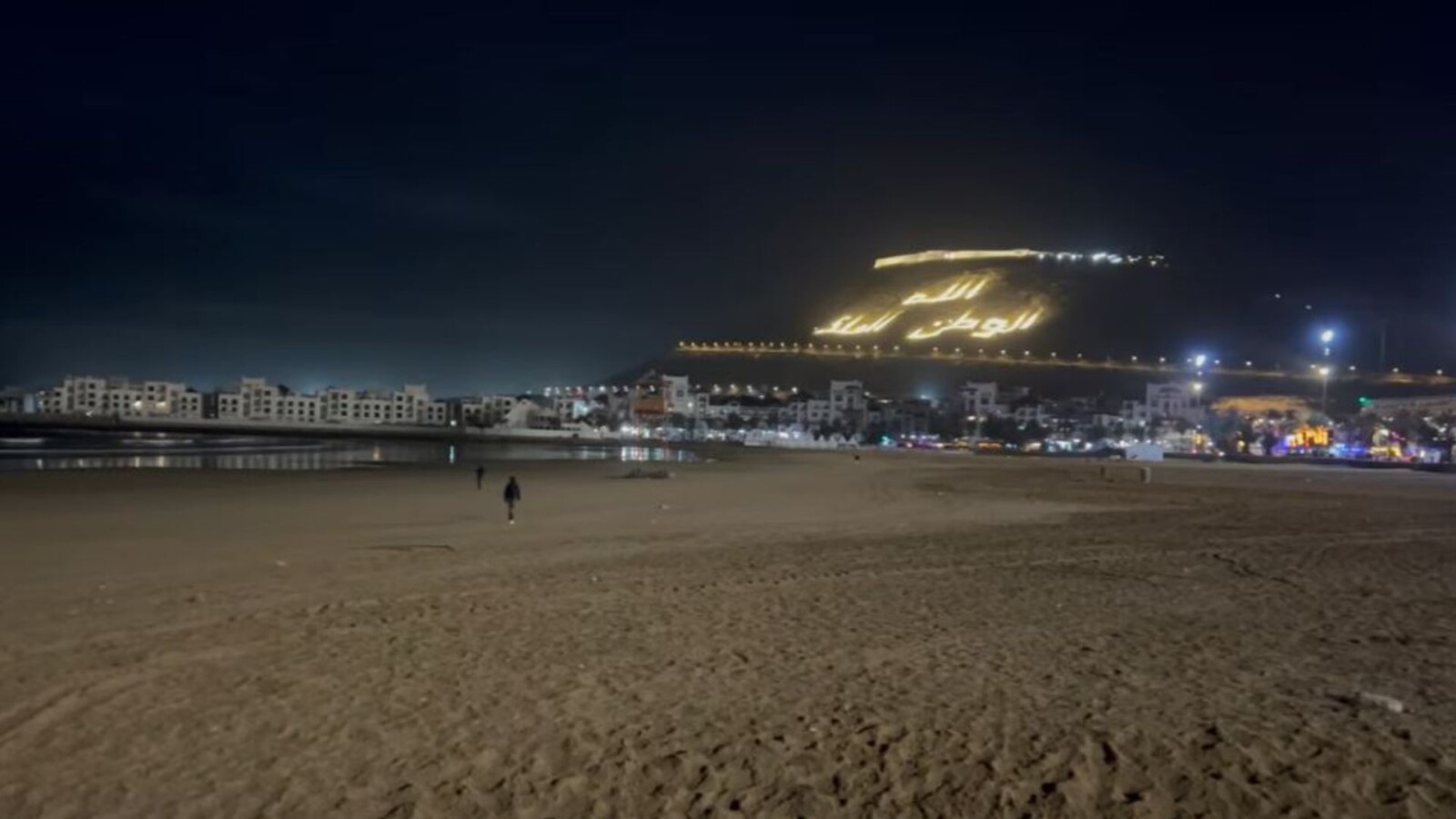 Night view of Agadir beach with a nearly empty stretch of sand, illuminated seafront buildings, and the glowing hillside inscription “Allah, Al Watan, Al Malik” above the city