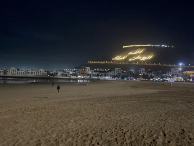 Night view of Agadir beach with a nearly empty stretch of sand, illuminated seafront buildings, and the glowing hillside inscription “Allah, Al Watan, Al Malik” above the city