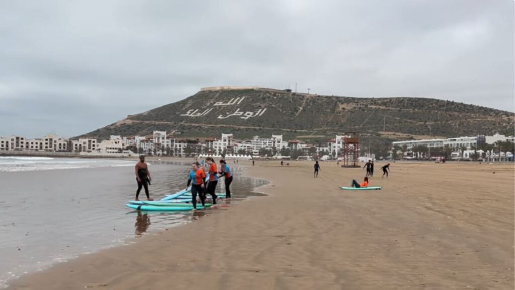 Surf students with boards on the wet sand of Agadir beach during a lesson, with the white hillside inscription “Allah, Al Watan, Al Malik” and seafront hotels visible in the background under a cloudy sky