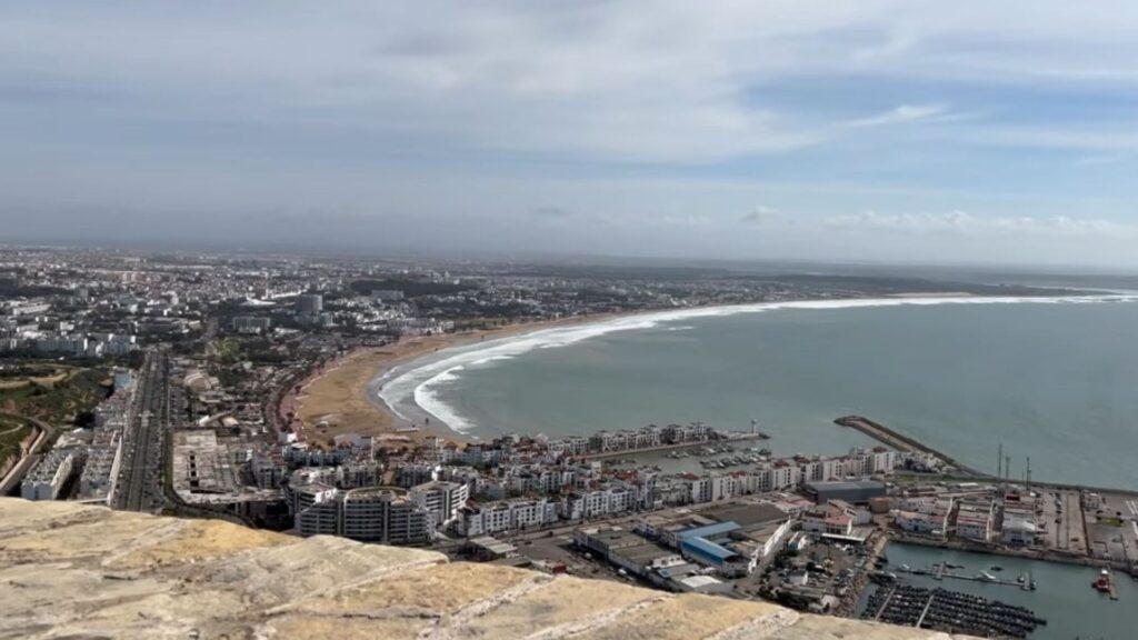 Panoramic daytime view over Agadir from the hilltop kasbah, showing the sweeping crescent-shaped beach, marina, and dense cityscape along the Atlantic coastline