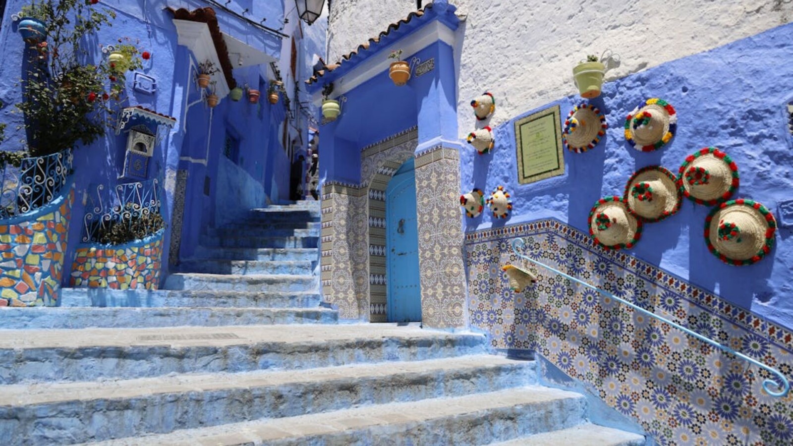 Sunlit alley in Chefchaouen featuring vivid blue-painted walls, stone steps, decorative Moroccan tiles, hanging flower pots, and colorful woven hats mounted on the wall, capturing the iconic charm of Morocco’s Blue City.