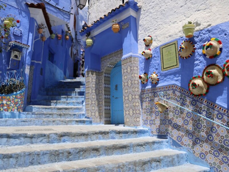 Sunlit alley in Chefchaouen featuring vivid blue-painted walls, stone steps, decorative Moroccan tiles, hanging flower pots, and colorful woven hats mounted on the wall, capturing the iconic charm of Morocco’s Blue City.