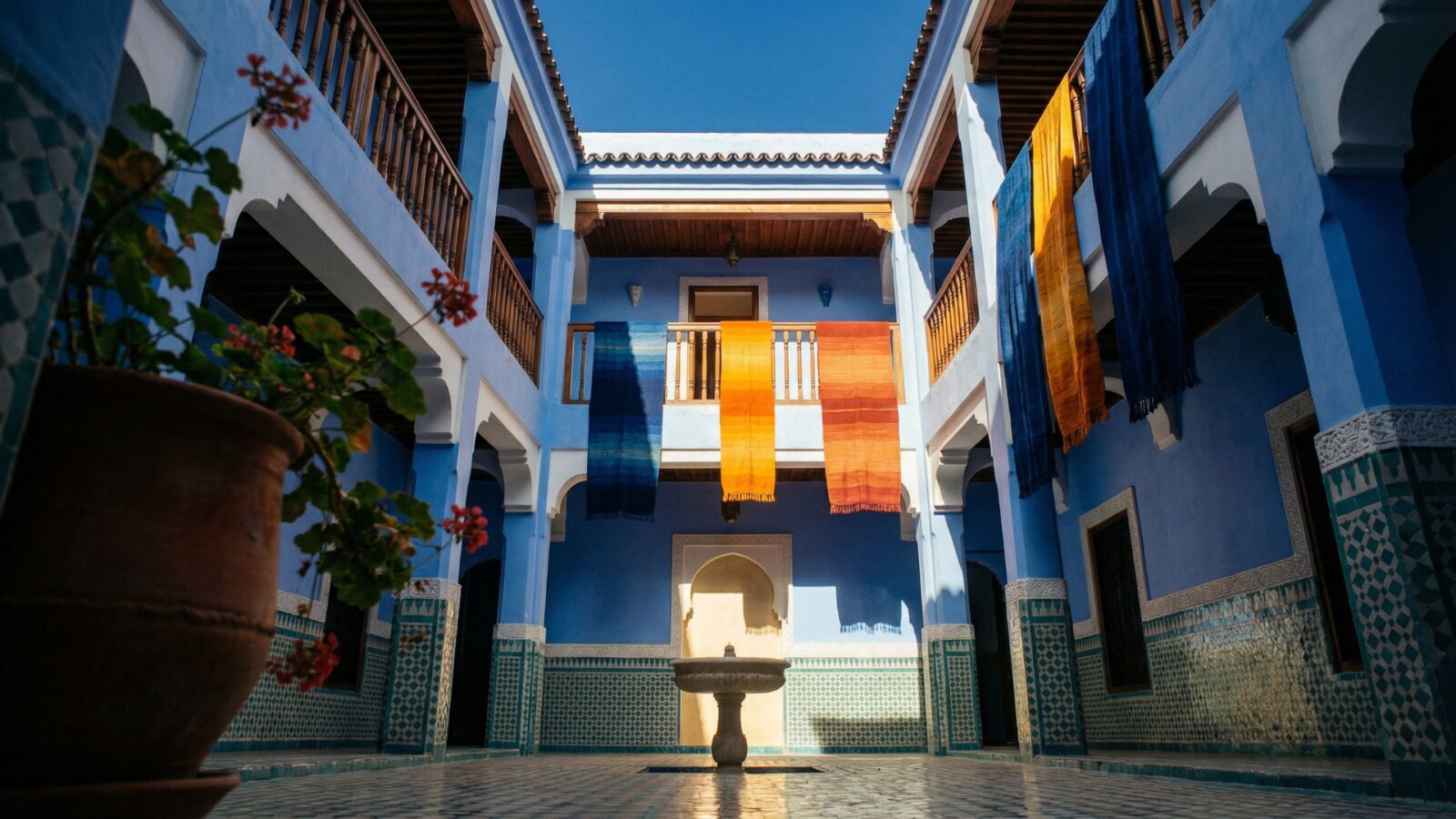 Interior courtyard of a traditional blue riad in Chefchaouen medina with zellige tilework, hanging textiles, and open sky above, Morocco