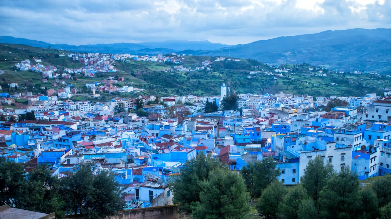 Panoramic view of Chefchaouen, a Moroccan town with blue-painted houses, green hills, and mountains in the background under a cloudy sky