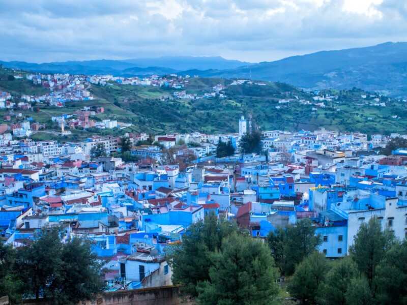 Panoramic view of Chefchaouen, a Moroccan town with blue-painted houses, green hills, and mountains in the background under a cloudy sky