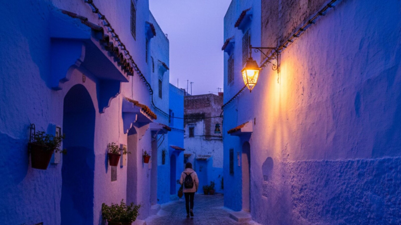 Solo traveler walking through a narrow blue alleyway in Chefchaouen medina at dusk with a traditional lantern casting warm amber light on wet cobblestones, Morocco