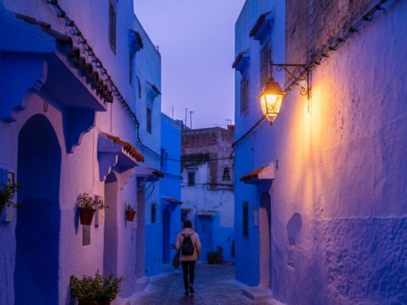 Solo traveler walking through a narrow blue alleyway in Chefchaouen medina at dusk with a traditional lantern casting warm amber light on wet cobblestones, Morocco