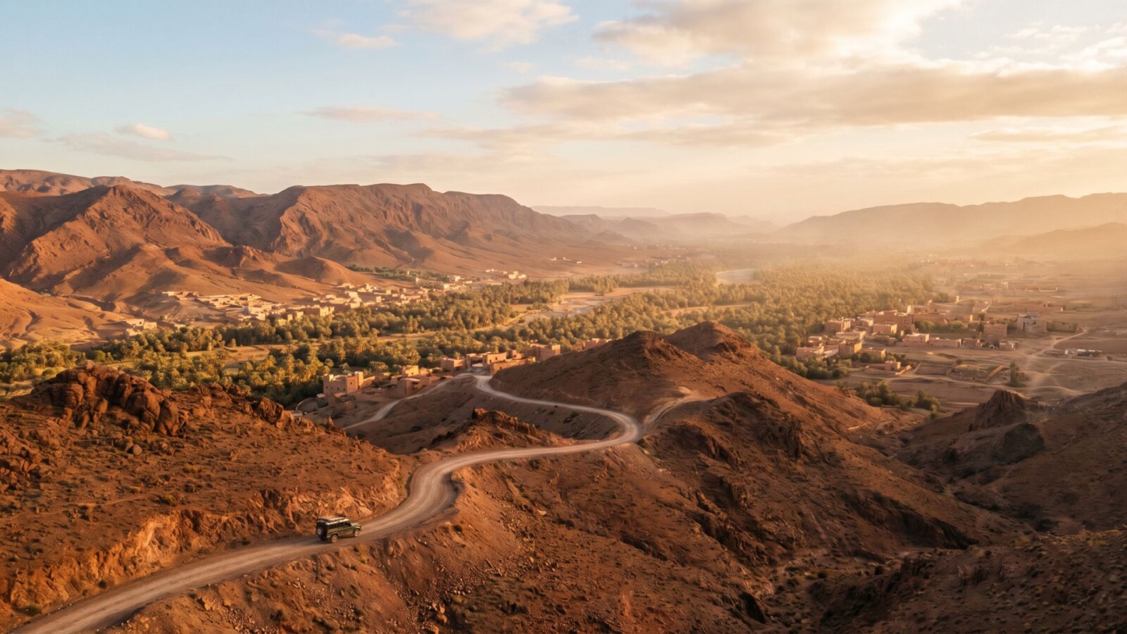 Winding High Atlas mountain road at golden hour looking south toward the Draa Valley in Morocco, with Berber villages and date palms visible in the distance below.