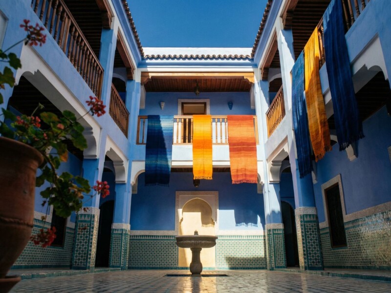 Interior courtyard of a traditional blue riad in Chefchaouen medina with zellige tilework, hanging textiles, and open sky above, Morocco