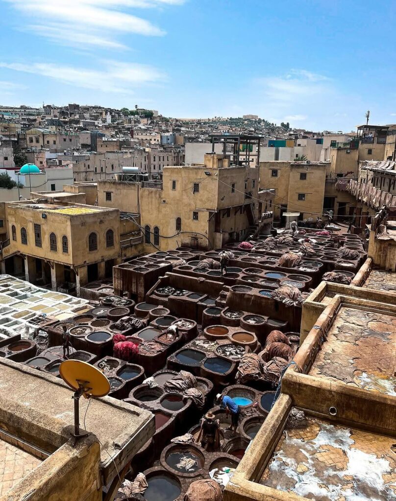 View over the traditional tanneries of Fes, Morocco, showing rows of circular dye vats, workers, and the dense old-city buildings under a bright blue sky.