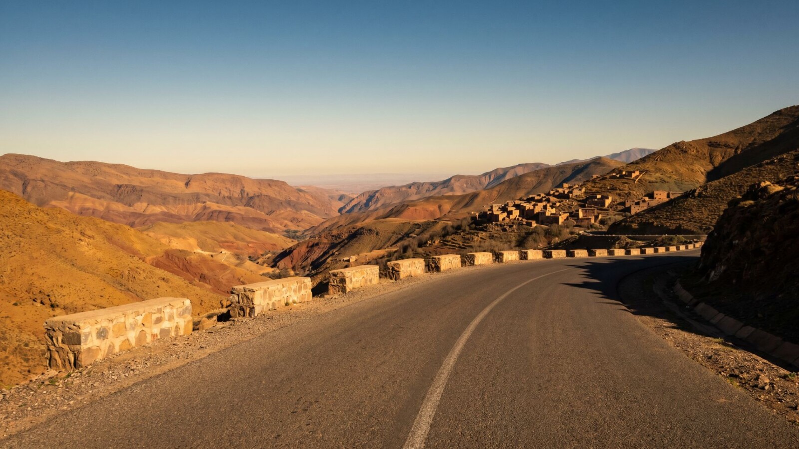 Empty mountain road winding through the High Atlas of Morocco at golden hour, with layered ochre peaks and a distant Berber village visible in the valley below.